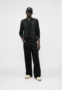 Μαύρο polo με φερμουάρ και κατακόρυφες ρίγες, συνδυασμένο με φαρδύ παντελόνι μαύρου χρώματος. Λευκά αθλητικά παπούτσια με καφέ σόλες. Το μαύρο καπέλο ολοκληρώνει την εμφάνιση.