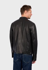 Chaqueta de cuero negro con una textura suave, un diseño estructurado, cuello alzado y cremalleras laterales. Combinada con unos vaqueros de mezclilla azul.