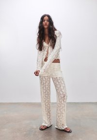 Spitzenoutfit mit einem kurz geschnittenen, gebundenen Oberteil mit langen Ärmeln und passenden weiten Hosen, beide in Weiß mit einem Blumendesign.