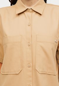 Το κοντομάνικο πουκάμισο διαθέτει κούμπωμα με κουμπιά μπροστά, γιακά, δύο τσέπες στο στήθος και ένα μικρό μεταλλικό λογότυπο σε μία από τις τσέπες.