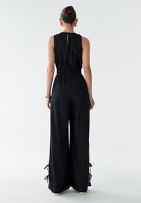 Mono negro con cintura entallada, piernas anchas y detalles ajustables con lazos en los puños. La parte trasera presenta una pequeña abertura tipo ojal.