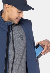 Marinefarbene Zip-Jacke über einem grauen gemusterten Sweatshirt, in die eine blaue Smartphone in die Tasche der Jacke gesteckt wird.