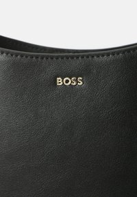 Sac en cuir noir avec une surface texturée. Logo "BOSS" doré affiché en évidence. Lignes épurées et design minimaliste.