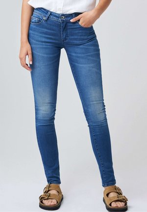 Kobieta w dopasowanych niebieskich dżinsach z przetarciami na udach i brązowych sandałach z klamrą, z jedną ręką w kieszeni, stoi na jednolitym tle.