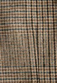 Brūnas un bēšas krāsas rūtainā auduma ar teksturētu virsmu. Audumā ir režģa formas laukumi, kuros sīkas melnas līnijas piešķir kontrastu.