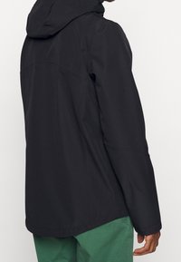 Černá nepromokavá bunda s kapucí, vybavená hladkou texturou a přiléhavým střihem, zobrazená vzadu s zelenými kalhotami.