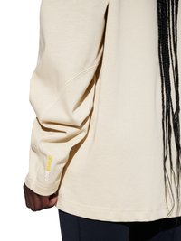 Osoba nosząca beżową bluzę z długim rękawem z logo „CORDURA” na mankiecie oraz czarne spodnie, z długimi splecionymi włosami widocznymi po prawej stronie.