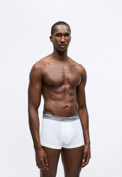 Fehér boxer alsónadrág, szürke márkás derékpánttal, szoros szabású és sima anyagú, bemutatva egy férfi modellen, sima háttér előtt.