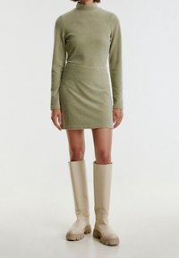 Robe longue à manches longues verte à col montant, silhouette ajustée, complétée par des bottes beiges clair jusqu'au genou avec des semelles texturées.