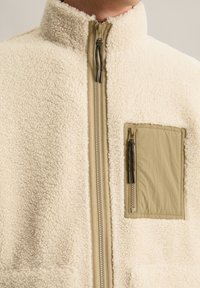 Crème kleurige fleece jas met een hoge kraag, voorzien van een beige rits aan de voorkant en een borstzak. Gestructureerde, zachte stof met een gestileerd ontwerp.