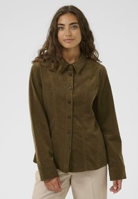 Woman with long curly brown hair wearing a fitted olive corduroy button-up shirt and beige pants on a white background.