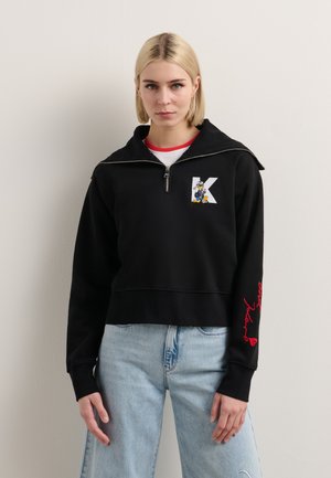 Black cropped hoodie with a zipper, featuring a white letter "K" and a colorful character design. Red text accents on the sleeve.