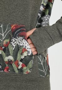 Sudadera gris con un diseño de bolsillo floral que presenta varios colores, incluyendo rojo y verde. Hecha de una tela suave con un acabado texturizado.