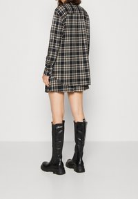 Abito camicia a quadri neri con maniche lunghe, caratterizzato da dettagli bianchi e marroni, abbinato a stivali neri lucidi fino al ginocchio e con orlo leggermente svasato.