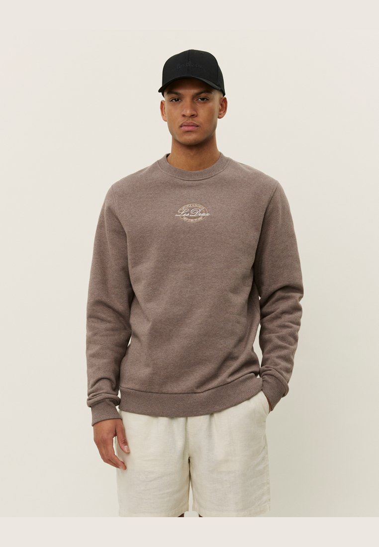 Brauner Sweatshirt mit Rundhalsausschnitt und gerippten Bündchen, mit einem kleinen grafischen Druck auf der Brust. Kombiniert mit hellen Shorts und einer schwarzen Kappe.