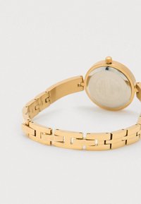 Gold metal watch with a round case, smooth link bracelet, and a clear glass face, featuring engraved brand names on the links.