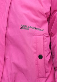 Jachetă roz aprins, cu o textură netedă, dotată cu un buzunar lateral și un buton negru, având imprimat logo-ul "KARL LAGERFELD JEANS" în litere închise.