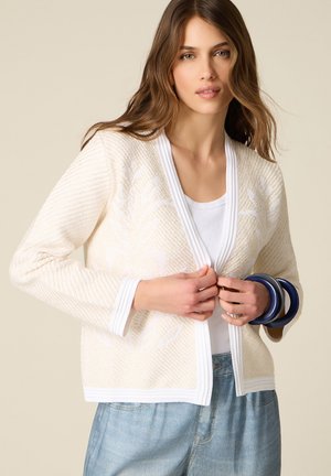 Donna che indossa un cardigan crema testurizzato sopra una camicia bianca, jeans azzurri chiari e tiene braccialetti blu scuro al polso.