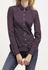 Chemise boutonnée bordeaux à pois blancs, à manches longues, avec col et coupe ajustée, présentant une texture lisse et un style classique.
