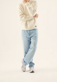 Cremfarbener, gerippter Hoodie mit Fronttasche, kombiniert mit hellblauen, lockeren Jeans und grauen Sportschuhen, die einen entspannten, lässigen Look erzeugen.