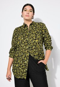 Camisa de manga larga, abotonada, en verde oliva con estampado de leopardo negro y amarillo. Presenta un collar y mangas enrolladas. Llevada con pantalones negros.