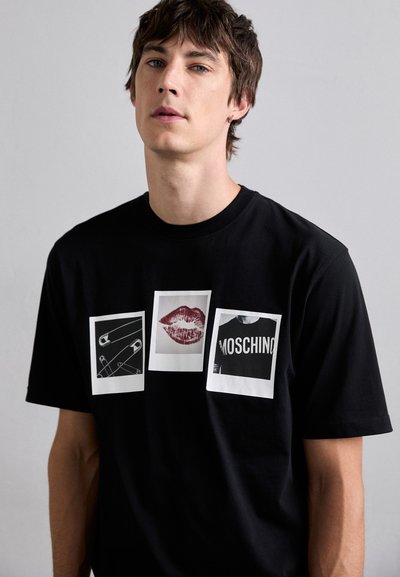 T-shirt en coton noir avec trois impressions graphiques : une épingle de sûreté, une marque de rouge à lèvres rouge et le texte "MOSCHINO" en blanc sur fond noir.