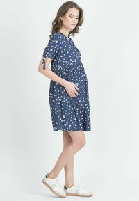 Marineblaue, bedruckte Umstandsbluse mit kurzen Ärmeln, gerafftem Taillengürtel und seitlichen Bändern. Kombiniert mit weißen Sneakern, die grüne und gelbe Akzente aufweisen.