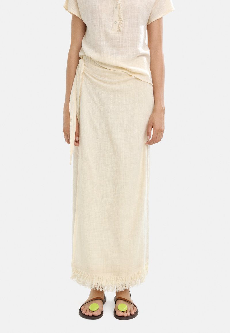 Luźna bluzka z kremowego lnu z listwą guzikową oraz długa, dopasowana spódnica z frędzlami na brzegu. Nosi się z płaskimi, brązowymi sandałami z zielonymi akcentami.