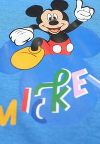 Tissu bleu avec un motif d'un personnage de dessin animé ayant une tête noire, un short rouge à pois blancs et des chaussures jaunes, accompagné de texte coloré.