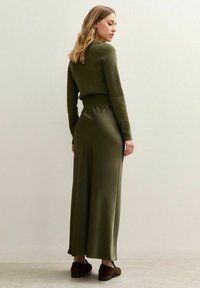 Femme aux cheveux blonds mi-longs portant un haut à manches longues vert olive et une jupe longue assortie, se tenant contre un mur uni.