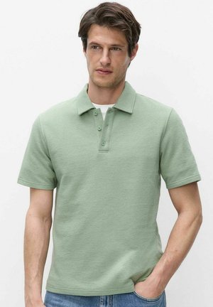Mand iført lysegrøn kortærmet polo shirt med tre knapper og blå jeans, står med den ene hånd i lommen mod hvid baggrund.