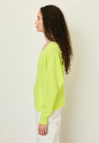 Neon gele oversized trui met een wijde halslijn, lange mouwen en een zachte textuur, gecombineerd met witte high-waisted pantalons.
