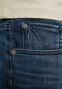 Vaqueros de mezclilla azul oscuro con un bolsillo frontal con herrajes de metal, costuras decorativas y una pequeña etiqueta encima del bolsillo.