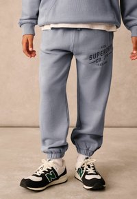 Lyseblå sweatpants med en manchet nederst, der har et sort trykt logo på låret, sammen med sorte og hvide sneakers.