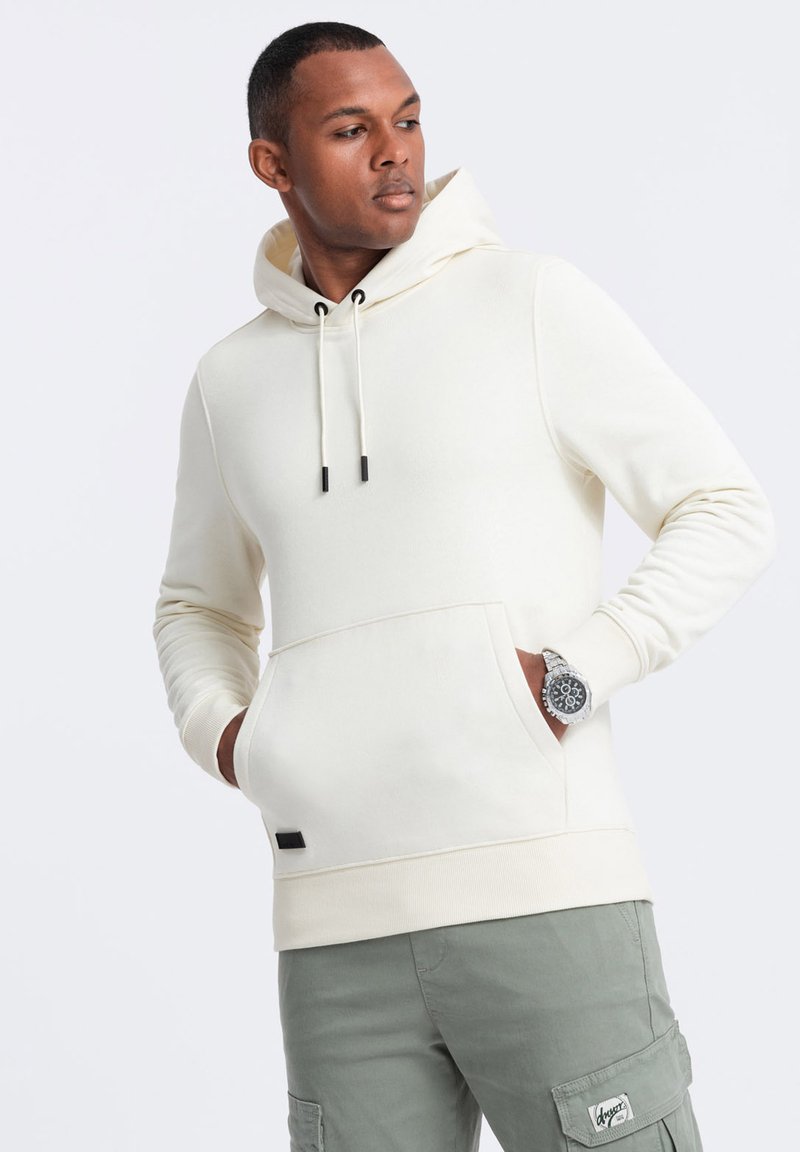 Felpa crema con cappuccio e coulisse, tasca frontale e polsini a coste. Abbinata a pantaloni cargo verdi, presenta un dettaglio con etichetta nera.