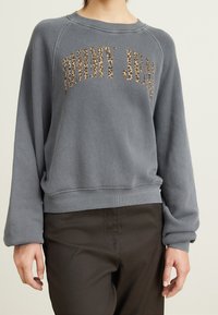 Kvinna som bär en grå sweatshirt med "TOMMY JEANS" i leopardsmönster, tillsammans med mörka byxor, poserande mot en neutral bakgrund.