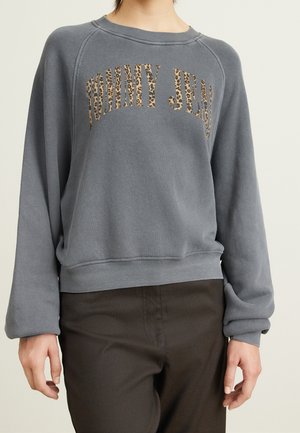 Frau trägt ein graues Sweatshirt mit "TOMMY JEANS" im Leopardenmuster, kombiniert mit dunklen Hosen, posiert vor einem neutralen Hintergrund.