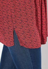 Rotes Hemd mit einem schwarzen wirbelnden Muster, mit seitlichen Schlitzen, kombiniert mit dunkelblauen Jeans, die eine glatte Textur und eine taillierte Silhouette aufweisen.
