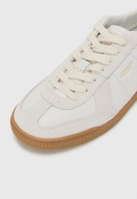 Hvit lærsneaker med semsket detaljer, flate lisser og gummisåle. Har stiftede detaljer og en gulllogo på siden.