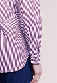 Röd och vitrandig långärmad skjorta med knappmanschett, med rundad nederkant och diskret logostickning på ärmen.