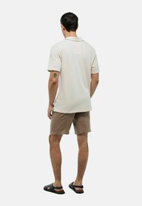 Polo shirt crème, à manches courtes, associé à un short beige. Le modèle porte des sandales noires. Le tissu semble lisse avec une coupe décontractée.