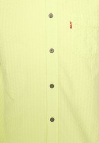 Jasnożółta koszula na guziki, teksturowana tkanina z małą kieszonką na piersi. Posiada pięć brązowych guzików oraz czerwoną metkę z logo na kieszeni.