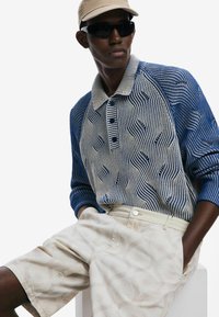 Blau-beiger Strick-Polo mit welligen Mustern, drei Knöpfen und langen Ärmeln; kombiniert mit beigen Shorts mit einem dezenten Muster.