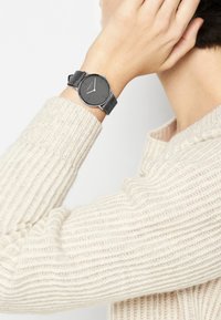 Runde Uhr mit grauem Ziffernblatt und minimalistischen silbernen Elementen. Schwarzes Lederarmband. Am Handgelenk getragen, im Kontrast zu einem cremefarbenen Strickpullover.