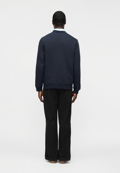 Άνδρας που στέκεται με την πλάτη γυρισμένη, φοράει μπλε σκούρο φούτερ, μαύρο παντελόνι και καφέ παπούτσια, μπροστά από απλό λευκό φόντο.