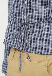 Κουστούμι μπλε και λευκό καρό πουκάμισο με κουμπιά, δεμένο σε φιόγκο στη μέση, συνδυασμένο με μπεζ παντελόνι ψηλ�όμεσο με κούμπωμα κουμπιού.