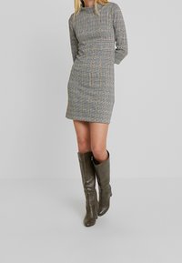 Abito grigio a quadri con maniche lunghe, una forma aderente e una texture liscia. Abbinato a stivali verdi al ginocchio con finitura in finta pelle.