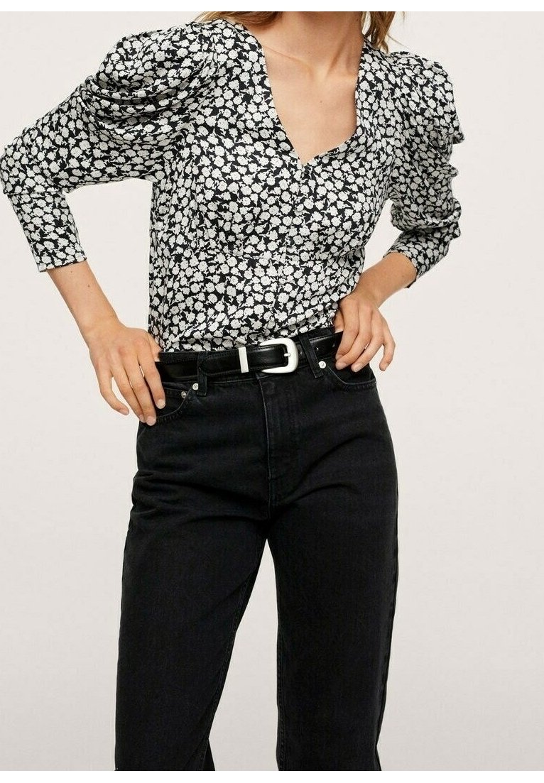 Femme portant un chemisier noir et blanc à motifs floraux avec des manches bouffantes, rentré dans un jean noir taille haute avec une ceinture noire.