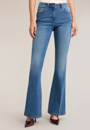Kvinde iført medium blå flared jeans og bordeaux spidse hæle, stående mod en neutral baggrund.