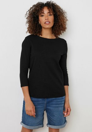 Femme aux cheveux bouclés portant un haut noir à manches trois-quarts et un short en jean bleu retroussé sur un fond uni.